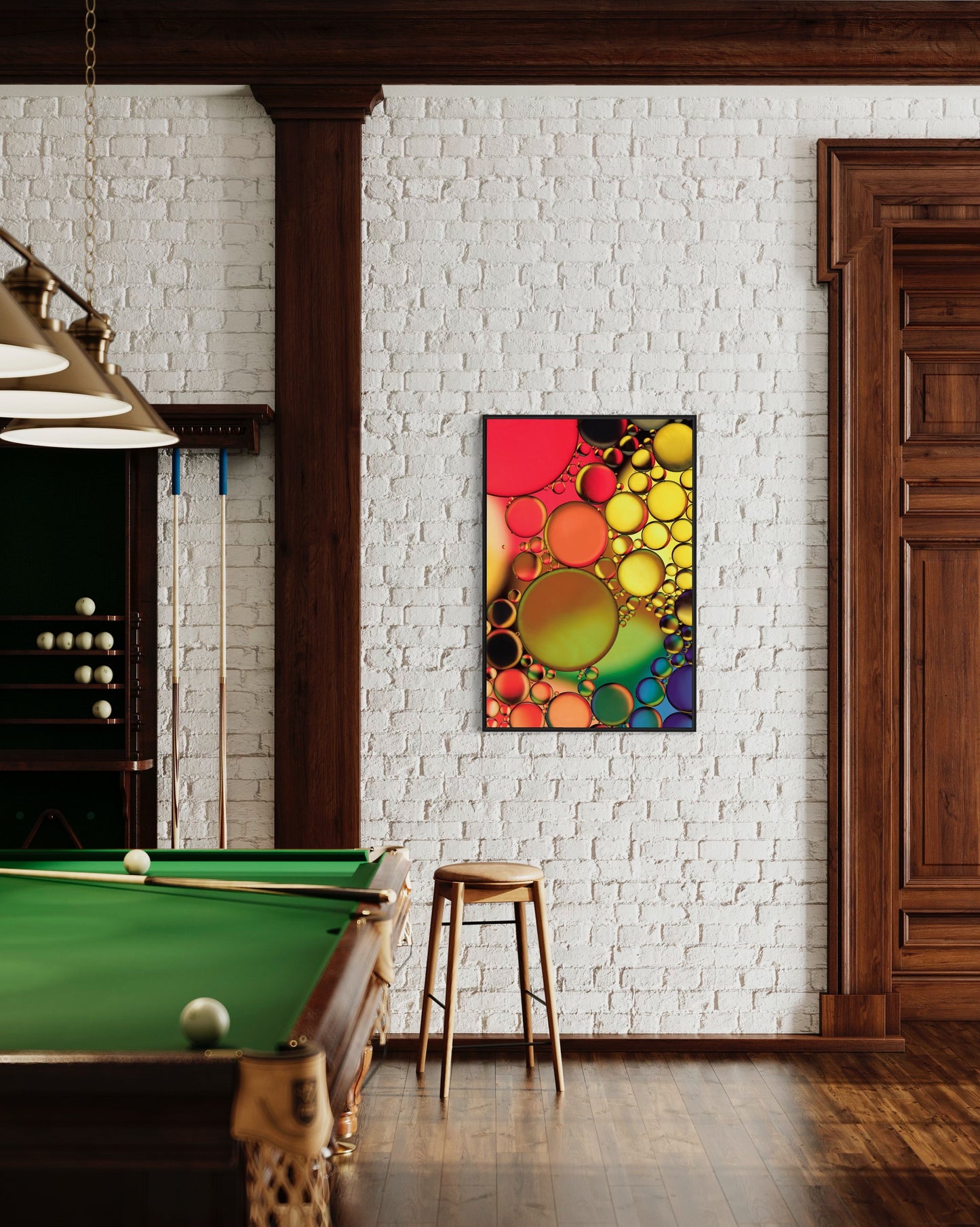 Abstract photograph titled "Jewels" featuring an interplay of oil and water in vibrant orange, yellow, green, blue, and purple hues, printed on high-grade photo paper and mounted under acrylic for depth and clarity. The predominant elements of this image are circular forms and vibrant hues. The image shows the print hanging on a white brick wall in a game room with pool table.