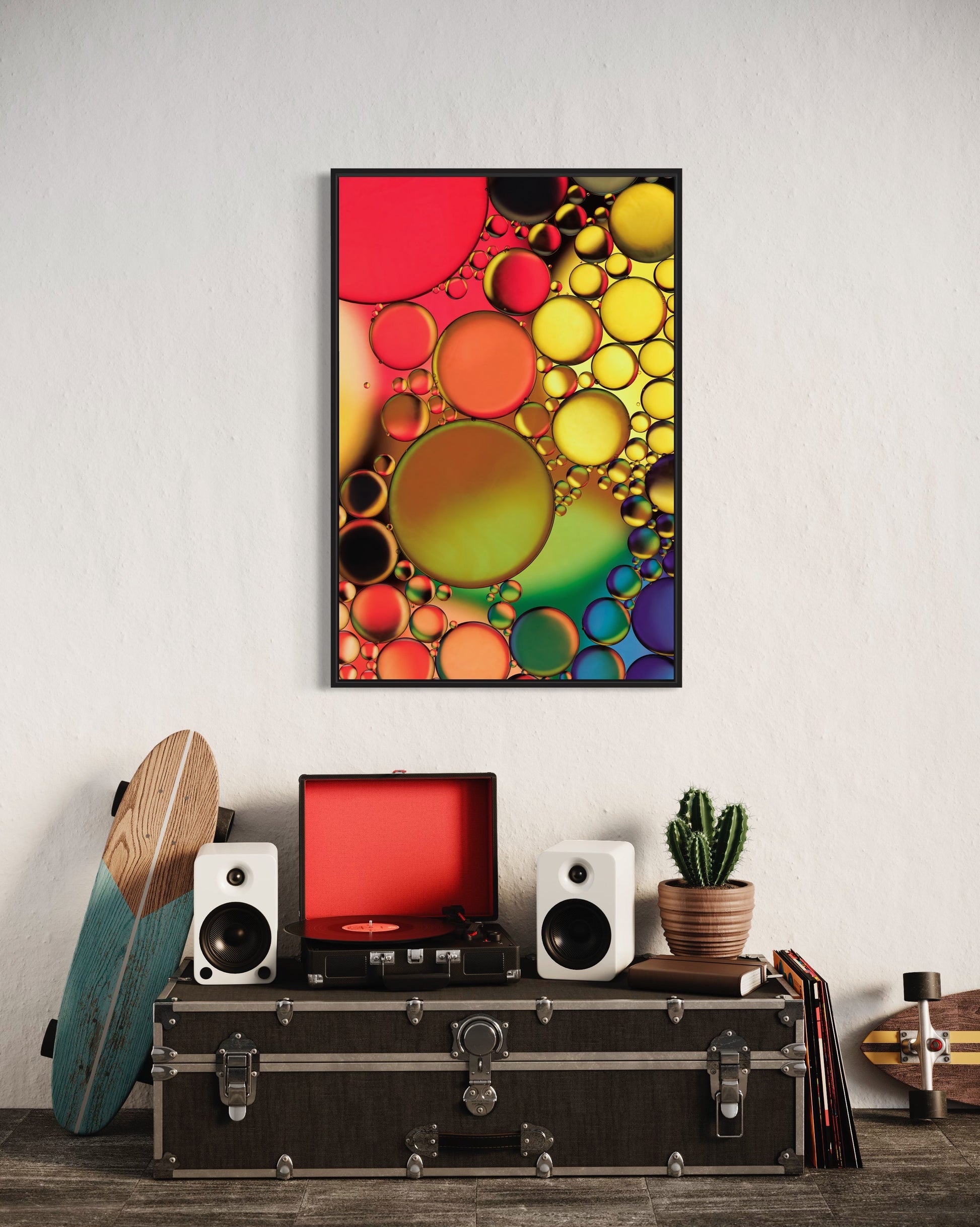 Abstract photograph titled "Jewels" featuring an interplay of oil and water in vibrant orange, yellow, green, blue, and purple hues, printed on high-grade photo paper and mounted under acrylic for depth and clarity. The predominant elements of this image are circular forms and vibrant hues.  The image shows the print hanging over  a trunk displaying a record player, plant and skateboard in a casual manner.